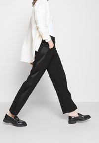 Zwarte broek met wijde pijpen, satijnen accenten, een getailleerde pasvorm en zakken. Gepaarde met een witte top en zwarte loafers.