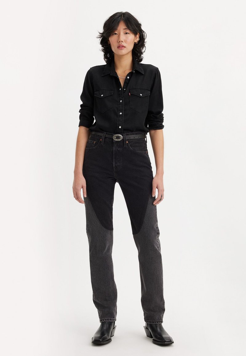 Camisa de mezclilla negra con cierre de botones y dos bolsillos en el pecho. Jeans de cintura alta en negro y gris con paneles en contraste. Botines negros.