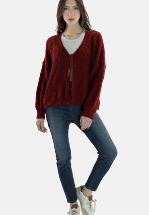 Cárdigan rojo texturizado con escote en V profundo y cierre de lazo, combinado con jeans azules y zapatillas blancas. Presenta mangas sueltas y extragrandes.
