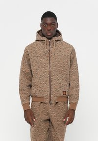 Bruine hoodie met een gestructureerd vlekkenpatroon, ritssluiting, koordjes voor de capuchon en zijzakken. Gemaakt van zacht materiaal met ribgebreide manchetten.