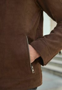 Giacca in suede marrone con tasca con zip, linee pulite e una texture morbida. La vestibilità è rilassata, con un colletto casual e dettagli alle maniche.