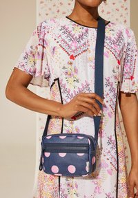 Jūras spēku krāsas plecu soma ar rozā punktiņiem, ar rāvējslēdzēja kabatām un struktūru. Siksna ir regulējama, ar izturīgu dizainu.