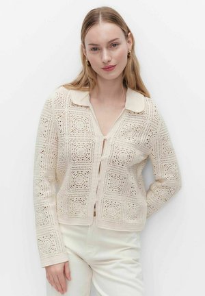 Ung kvinde iført cremefarvet hæklejakke med krave og hvide bukser, stående mod en ensfarvet hvid baggrund.