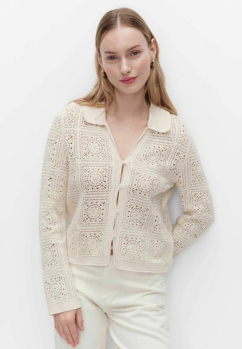 Jonge vrouw draagt een crème kleurige gehaakte cardigan met kraag en witte broek, staand tegen een effen witte achtergrond.