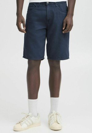 Mand iført marineblå knælange shorts, hvide crew-sokker og hvide Adidas-sneakers med lysegrå striber, stående på en ensfarvet baggrund.