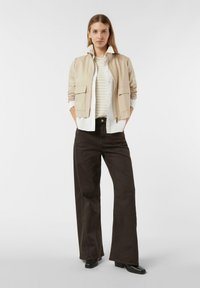 Beżowa kurtka na zamek z dwoma przednimi kieszeniami, nałożona na biało-beżową koszulę w paski, połączona z ciemnobrązowymi, wysokim stanem spodniami i czarnymi butami.