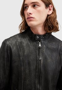 Giacca di pelle nera con colletto alto, cuciture ben visibili e chiusura frontale con zip. Il materiale ha una finitura opaca con un aspetto texture.