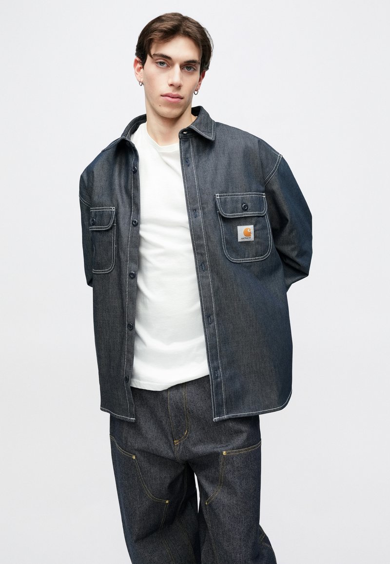 Mørk denim overshirt med knaplukning foran, brystlommer og kontrastsyning, båret over en hvid t-shirt og mørke denimbukser.