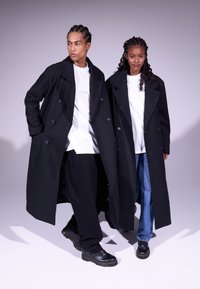 Manteaux noirs à double boutonnage avec de grands boutons, portés sur des chemises blanches et des pantalons sombres. Des mannequins se tiennent côte à côte sur un fond gris clair.
