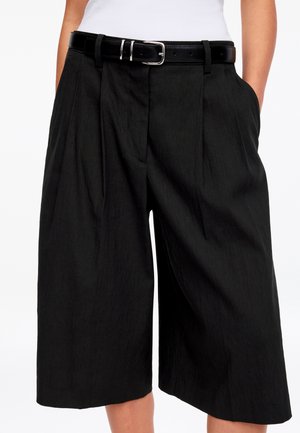 Sorte shorts med højt talje og vide ben, med folder foran, båret med sort bælte og hvid top stoppet ned, hænderne i lommerne.