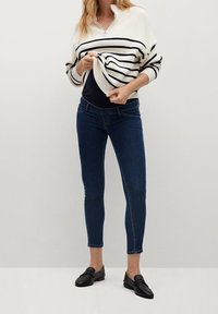 Mujer vistiendo jeans ajustados oscuros, mocasines negros y un suéter blanco con rayas horizontales negras, levantando ligeramente el suéter.