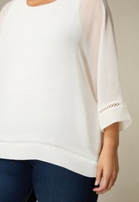 Donna che indossa una camicetta bianca trasparente con bordo in pizzo circolare sulle maniche e sull'orlo, abbinata a jeans in denim blu scuro.