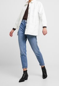 Chaqueta de mezclilla blanca con botones y bolsillos, combinada con jeans de cintura alta azul y botas negras hasta el tobillo. Top a rayas visible debajo.