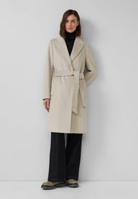 Beige Wollmischungscoat mit tailliertem Schnitt, Reverskragen, langen Ärmeln und seitlichen Taschen, kombiniert mit schwarzen weiten Hosen und klobigen Stiefeln.