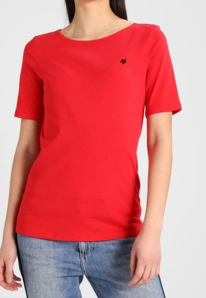 Kobieta w jednolitej czerwonej koszulce z krótkim rękawem z małym czarnym logo na piersi, w połączeniu z jasnoniebieskimi dżinsami.