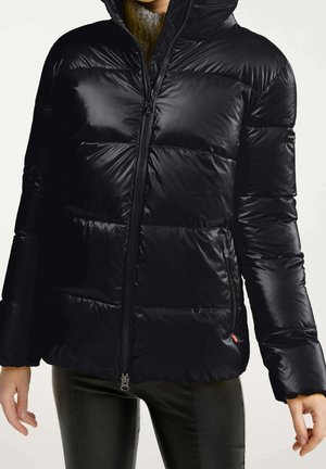 Femme portant une doudoune noire brillante avec fermeture éclair devant et poches zippées, associée à un pantalon noir et un pull col roulé gris.