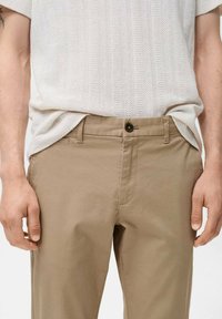 Chino beige con una texture liscia e opaca, dotati di un bottone frontale, passanti per la cintura e tasche laterali. Abbinati a una maglia leggera e texturizzata.