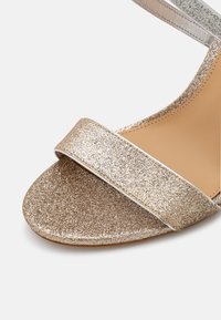 Gros plan d'une sandale dorée pailletée à bout ouvert avec une bride unique sur les orteils et une semelle intérieure beige lisse.