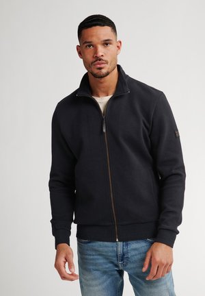 Zwarte zip-up jas met een textuurpatroon, geribde manchetten en zoom, met een klein logopatch op de mouw, gedragen over een beige shirt.