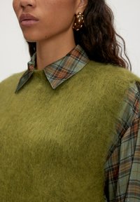 Groene, fuzzy gebreide vest over een bruin-groene geruite shirt met een spitse kraag. Gouden accenten in de oorbellen maken het geheel af.