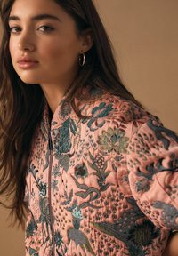 Gewatteerd jack van roze stof, met ingewikkeld borduurwerk van bloemen en vogels in groene en blauwe tinten, voorzien van een rits en een hoge kraag.