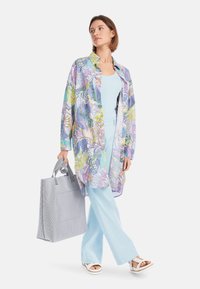 Bunte lange Shirtjacke mit floralem und Sushi-Muster, kombiniert mit einem hellblauen Oberteil und Hose. Graue gestreifte Tragetasche. Weiße Sandalen.