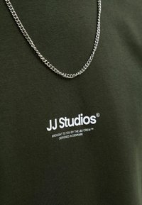 Camiseta verde oliva con cuello redondo, hecha de una tela suave. Presenta un logo de texto blanco "JJ Studios" y un collar de cadena plateada brillante.