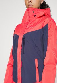 Röd och marinblå regnjacka med hög krage, dragkedjeförsedd bröstficka, justerbar dragsko-huva och slätt, vattenavvisande tyg.