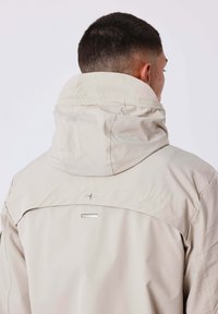 Uomo di spalle che indossa una giacca beige chiaro con colletto alto e cappuccio, che mostra un piccolo logo metallico del marchio sulla parte superiore della schiena.