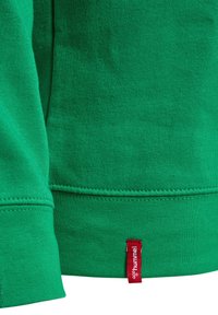 Groene sweatshirtmouw en zoom met een klein rood stoffen label met de tekst "hummel" dicht bij de onderrand vastgenaaid.
