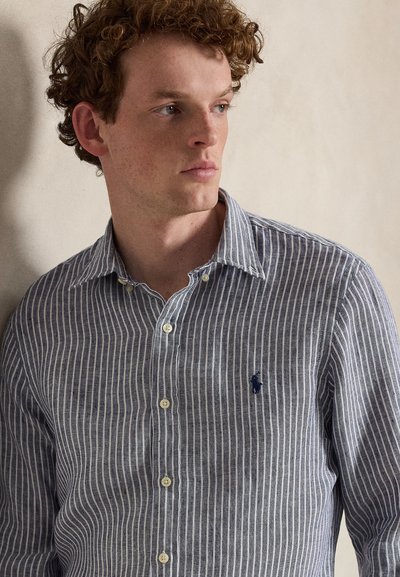 Νεαρός με σγουρά μαλλιά που φοράει μπλε και λευκό πουκάμισο με κάθετες ρίγες κοιτάζει προς τα πλάγια σε απλό φόντο.