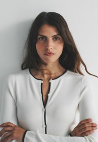 Cardigan alb cu model nervurat, margini negre și închidere cu nasturi, cu decolteu rotund. Textură moale și mâneci lungi vizibile.