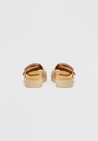 Beigefarbene Slides mit einem erhöhten, strukturierten Akzent am Riemen und einer cremefarbenen Sohle. Das Design ist offen mit niedrigem Absatz.