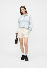 Νέα γυναίκα με φωτεινό μπλε ριγέ μακρυμάνικο πουκάμισο, κρεμ σορτς, μαύρα αθλητικά παπούτσια, λευκές κάλτσες, κρατώντας μαύρη τσάντα χειρός, στέκεται σε λευκό φόντο.