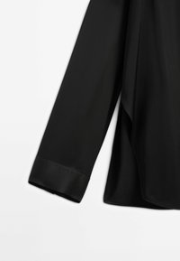 Manche de chemise noire à manches longues avec un large poignet, montrée sur un fond blanc, mettant en valeur la texture du tissu et les détails des coutures.