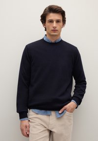 Giovane uomo in piedi contro un muro chiaro, indossa un pullover blu navy sopra una camicia azzurra e pantaloni beige, con una mano in tasca.