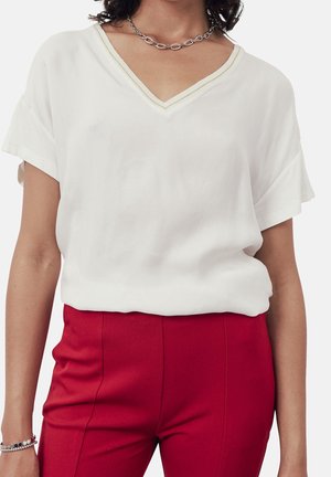 Nainen, joka on pukeutunut valkoiseen V-kaula-aukkotoppiin, jossa on lyhyet hihat, yhdistettynä korkeavyötäröisiin punaisiin housuihin, hopeiseen ketjukaulaketjuun ja rannekoruun.