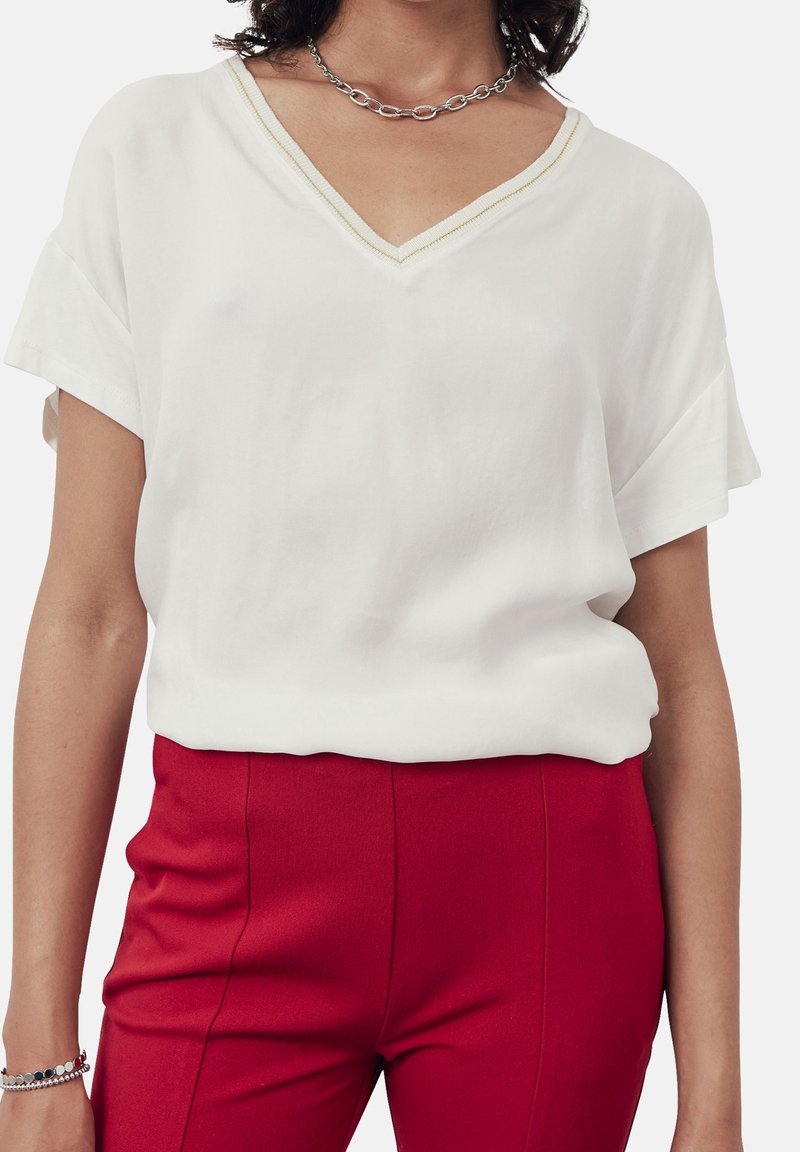Femme portant un haut blanc à col en V et manches courtes associé à un pantalon rouge taille haute, une chaîne en argent autour du cou et un bracelet.