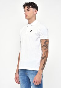 Polo shirt bianca in cotone con pattina a due bottoni e una piccola etichetta con logo sul petto. Maniche corte e vestibilità regolare. Indossata con jeans blu.