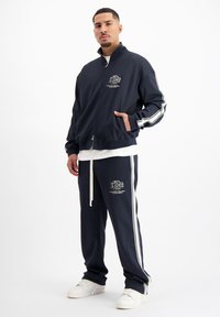 Marine trainingspak met een ritsjack en broek met aantrekkoord. Voorzien van witte strepen op de mouwen en zijkanten van de broek. Logo op het jack en de broek.