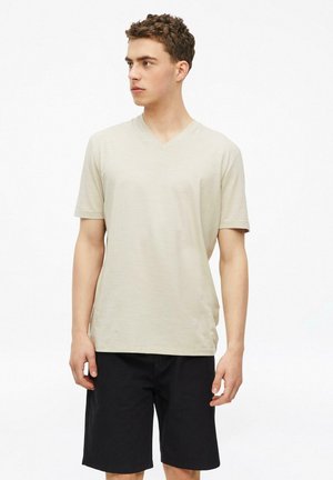 Młody mężczyzna z krótkimi kręconymi włosami, ubrany w beżowy T-shirt z dekoltem w kształcie litery V i czarne szorty, patrzący na bok na jednolitym białym tle.