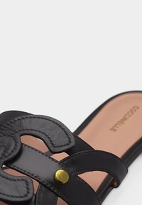 Sandale en cuir noir avec des brides décoratives découpées et un rivet en laiton sur une semelle intérieure beige.