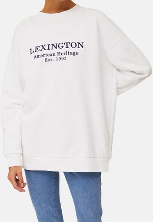 Valkoinen pyöreäkauluksinen collegepaita, jossa teksti "LEXINGTON American Heritage Est. 1993", yhdistetty sinisiin farkkuihin, oikea käsi lanteilla.
