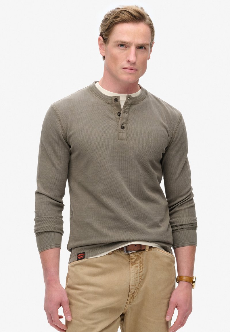Olijf groene shirt met lange mouwen in henley-stijl met drie knopen, geribbelde textuur en een crèmekleurige kraag. Draagt met een tan kleurige broek en een bruine riem.