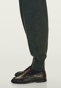 Benen in donkergroene tapered pantalon en zwarte leren loafers met geplooide details, gestyled over zwarte sokken tegen een lichte achtergrond.