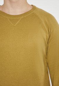 Mustársárga hosszú ujjú póló, kerek nyakkivágással, bordázott textúrával és raglán ujjakkal. A varrások mentén megerősített varrási részlet található.