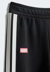 Pantalons de survêtement noirs avec deux rayures blanches sur les côtés et un patch rouge du logo Marvel sur le bas devant. Texture de tissu doux et côtelé.