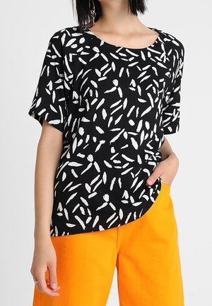 Vrouw draagt een zwart T-shirt met korte mouwen met een wit abstract penseelstrepenpatroon en feloranje wijde broekspijpen, hand in haar zak.