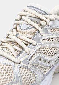 Baskets de sport beige clair avec une tige en mesh, accents argent métallisé et un col rembourré. Présente des lacets fins et une semelle texturée.