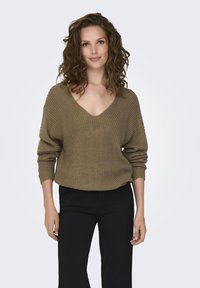 Khaki oversized trui van gebreid materiaal met een diepe V-hals, geribbelde mouwen en een relaxte pasvorm, gecombineerd met zwarte broeken. Zachte textuur en casual ontwerp.